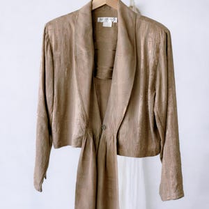 May include: A tan-colored jacket with a draped front and long sleeves hangs on a wooden hanger. The jacket has a textured, vertical striped pattern and a single button closure. The garment is displayed against a white background.