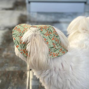May include: A light green fabric hat featuring a repeating pattern of orange berries and dark green leaves. The hat has a rounded crown and a short brim, designed to fit over the head with openings for ears, showcasing a botanical print.