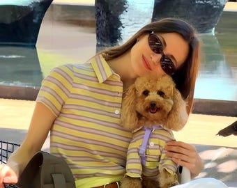 Puede incluir: Un polo amarillo con rayas rosas y grises, combinado con pantalones blancos y un cinturón marrón. Se sostiene un perro pequeño con una camisa a rayas a juego y un collar morado. La persona lleva gafas de sol.