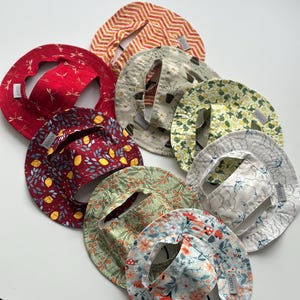 May include: A collection of eight circular fabric bibs with various patterns including red with dragonflies, orange chevron, white with daisies, yellow floral, lemon print, blue clouds, green foliage, and a multi-coloured floral print, each with an ODDY tag.