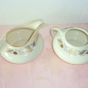 May include: A white creamer and sugar set with gold leaf detailing of leaves and branches. The set is on a pink surface.