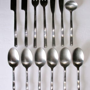May include: A set of thirteen stainless steel flatware pieces arranged in two rows. The top row includes two knives, two forks, two butter knives, and one small ladle. The bottom row consists of six matching spoons with a textured handle design.