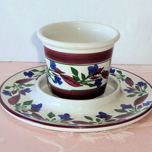 May include: A white ceramic cup and saucer set with a floral design. The cup and saucer are decorated with a burgundy band and blue and green floral accents.
