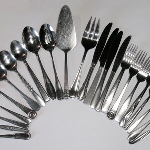 May include: An assortment of vintage silverware arranged in a semi-circle. The set includes spoons, forks, knives, and a cake server. The silverware is made of a shiny, silver-colored metal, reflecting the light.
