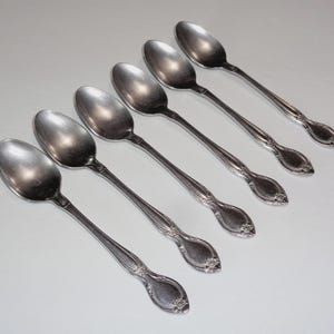 May include: Six silver spoons arranged diagonally across a white surface. Each spoon has a rounded bowl and a decorative handle with a floral design. The spoons are made of a shiny, reflective metal.