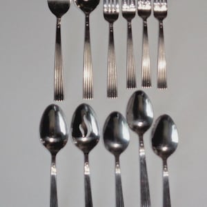 May include: A collection of stainless steel silverware, including forks and spoons. The utensils have a reflective, polished surface and a ribbed design on the handles. The arrangement includes various sizes of forks and spoons, suitable for a formal dining setting.