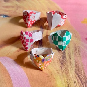 May include: Five origami heart rings in various colors and patterns. The rings are made of paper and have a white base. The patterns include red and white flowers, green and white checkered, and a multi-colored floral design.