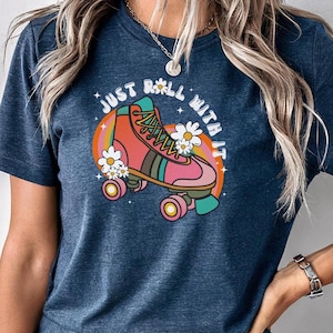 May include: A dark blue heathered t-shirt featuring a graphic of a pink and orange roller skate surrounded by white daisies and a rainbow arc. The text "JUST ROLL WITH IT" is printed in white letters above the roller skate illustration.