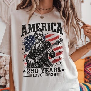 K&ouml;nnte beinhalten: Cremefarbenes T-Shirt mit einer Grafik von Abraham Lincoln, der Gitarre spielt, vor dem Hintergrund der amerikanischen Flagge. Der Text "AMERICA" steht &uuml;ber dem Bild, darunter "250 YEARS 1776-2026". Ein patriotisches Design.