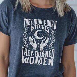 May include: Dark blue t-shirt featuring the white text "THEY DIDN'T BURN WITCHES THEY BURNED WOMEN". The design includes a crescent moon, stars, and hands. The shirt has a crew neck and short sleeves.