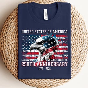 May include: Navy blue t-shirt featuring a graphic of an American flag and an eagle's head. The text reads "UNITED STATES OF AMERICA" and "250TH ANNIVERSARY 1776-2026". The shirt is made of a soft material and is suitable for casual wear.