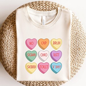 May include: A cream colored t-shirt with a design of nine candy hearts with different phrases on them. The phrases are "NO", "CAP", "BRUH", "SIGMA", "OHIO", "RIZZ", "SKIBIDI", "TOILET", and "V DAY".