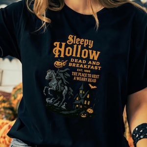 May include: Black t-shirt with a Halloween graphic. The design includes the text "Sleepy Hollow Dead and Breakfast" and an illustration of a headless horseman, a haunted house, and pumpkins. The shirt is a dark colour.
