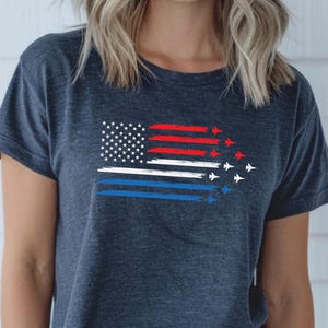 May include: A navy blue t-shirt featuring a patriotic design. The graphic includes a stylized American flag with red, white, and blue stripes, and a field of white stars. Small white jet plane silhouettes are trailing behind the stripes.