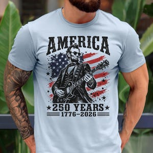 K&ouml;nnte beinhalten: Hellblaues T-Shirt mit einer Grafik von Abraham Lincoln, der Gitarre spielt, vor einer amerikanischen Flagge. Der Text auf dem Shirt lautet "AMERICA" und "250 YEARS 1776-2026".