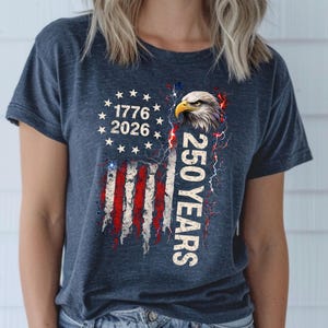 May include: Dark blue t-shirt featuring a distressed American flag design with the text "1776 2026" and "250 YEARS". The graphic includes an eagle head and stars.