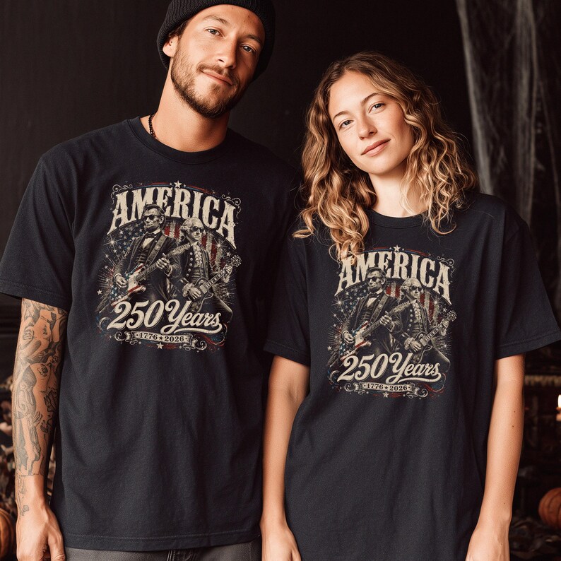 May include: Black t-shirts with a graphic design commemorating 250 years of America. The design features the word "America" above an illustration of two figures playing guitars, with the text "250 Years" and the years "1776-2026" below.