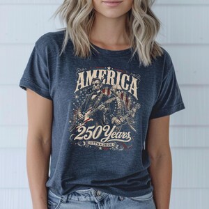 May include: Navy blue t-shirt featuring a graphic with the word "AMERICA" above an illustration of two figures playing guitars. The graphic also includes the text "250 Years" and the years "1776-2026".