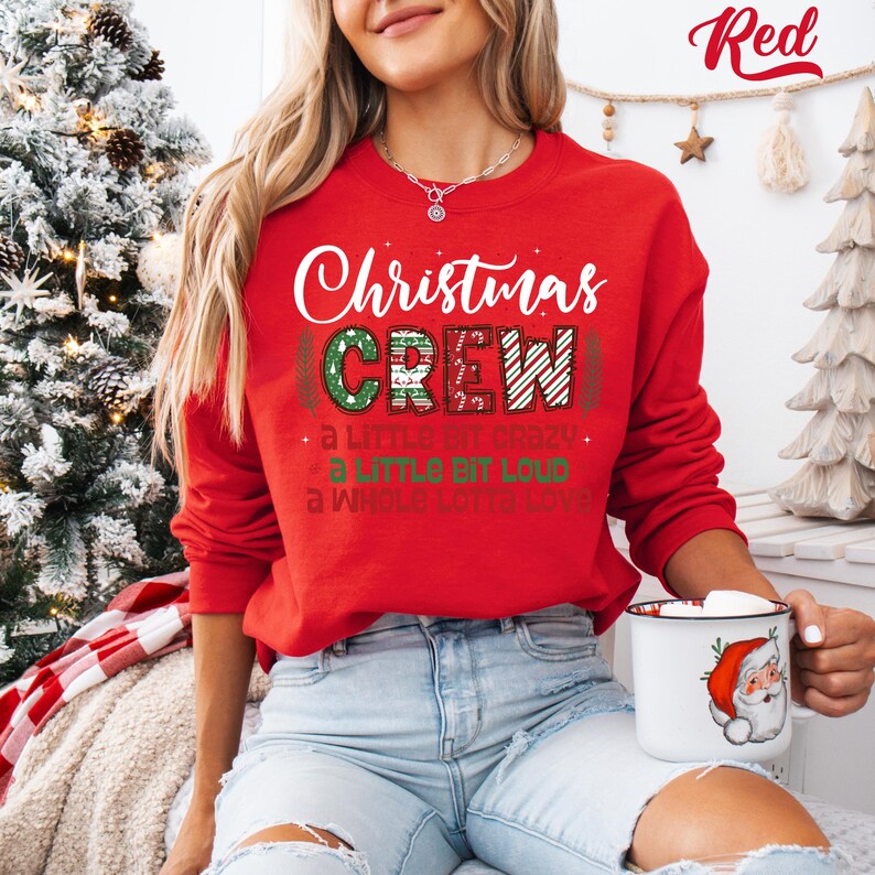 May include: A red Christmas sweatshirt with the words "Christmas CREW" in white and green. The sweatshirt has the phrases "a little bit crazy", "a little bit loud", and "a whole lotta love" printed below the main text. A white mug with a Santa Claus design is held in the hand.