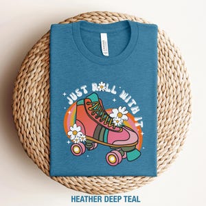 May include: A heather deep teal t-shirt featuring a graphic of a pink and orange roller skate with white daisies, a rainbow-colored arch, and the text "JUST ROLL WITH IT" in white letters. The shirt has a crew neckline and a white brand label.