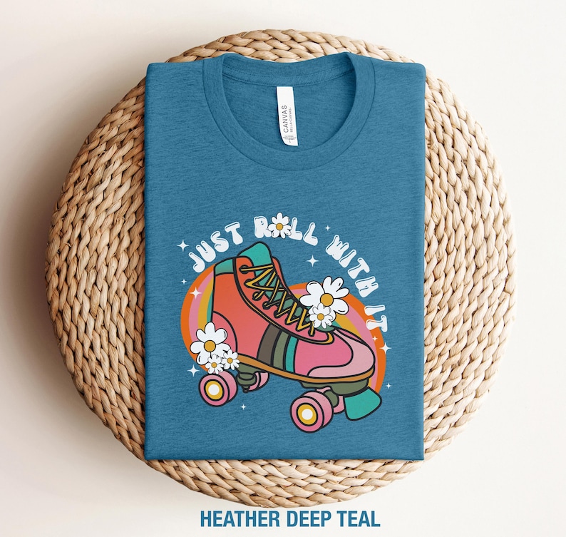 May include: A heather deep teal t-shirt featuring a graphic of a pink and orange roller skate with white daisies, a rainbow-colored arch, and the text "JUST ROLL WITH IT" in white letters. The shirt has a crew neckline and a white brand label.