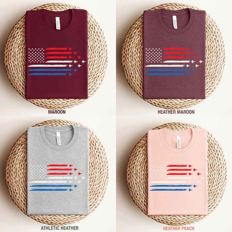 May include: Four t-shirts in maroon, heather maroon, athletic heather, and heather peach. Each shirt features a graphic of the American flag with red and blue stripes and white stars, with jets trailing behind. The shirts are displayed on woven placemats.