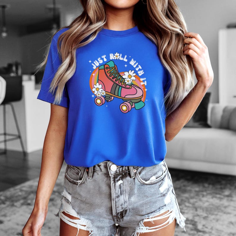 May include: A bright blue short-sleeve t-shirt featuring a graphic of a pink and teal roller skate surrounded by white daisies and a rainbow arc. The text "JUST ROLL WITH IT" is printed in a white arched font above the roller skate illustration.