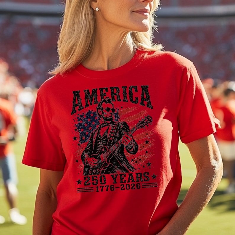 K&ouml;nnte beinhalten: Rotes T-Shirt mit dem Wort "AMERICA" in einer kr&auml;ftigen, verwitterten Schrift. Das Shirt zeigt eine Grafik von Abraham Lincoln, der Gitarre spielt, vor dem Hintergrund der amerikanischen Flagge und Sterne. Der Text "250 YEARS 1776-2026" ist ebenfalls aufgedruckt.