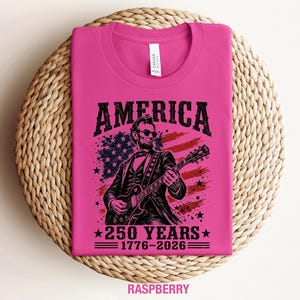 K&ouml;nnte beinhalten: Himbeerfarbenes T-Shirt mit einer Grafik von Abraham Lincoln, der vor einer amerikanischen Flagge Gitarre spielt. Das Wort "AMERICA" steht &uuml;ber dem Bild, darunter "250 YEARS 1776-2026". Das T-Shirt liegt gefaltet auf einer gewebten Oberfl&auml;che.