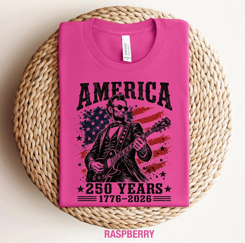 K&ouml;nnte beinhalten: Himbeerfarbenes T-Shirt mit einer Grafik von Abraham Lincoln, der vor einer amerikanischen Flagge Gitarre spielt. Das Wort "AMERICA" steht &uuml;ber dem Bild, darunter "250 YEARS 1776-2026". Das T-Shirt liegt gefaltet auf einer gewebten Oberfl&auml;che.