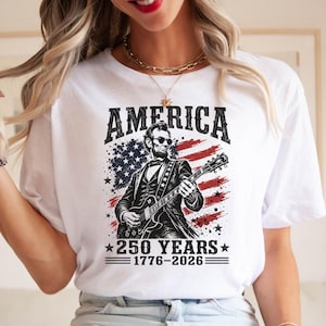 K&ouml;nnte beinhalten: Wei&szlig;es T-Shirt mit einer Grafik von Abraham Lincoln, der Gitarre vor einem amerikanischen Flaggen-Design spielt. Der Text "AMERICA" steht &uuml;ber dem Bild, darunter "250 YEARS 1776-2026".