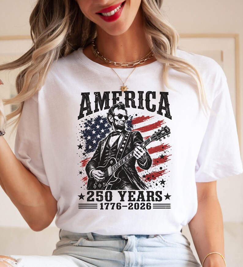 K&ouml;nnte beinhalten: Wei&szlig;es T-Shirt mit einer Grafik von Abraham Lincoln, der Gitarre vor einem amerikanischen Flaggen-Design spielt. Der Text "AMERICA" steht &uuml;ber dem Bild, darunter "250 YEARS 1776-2026".