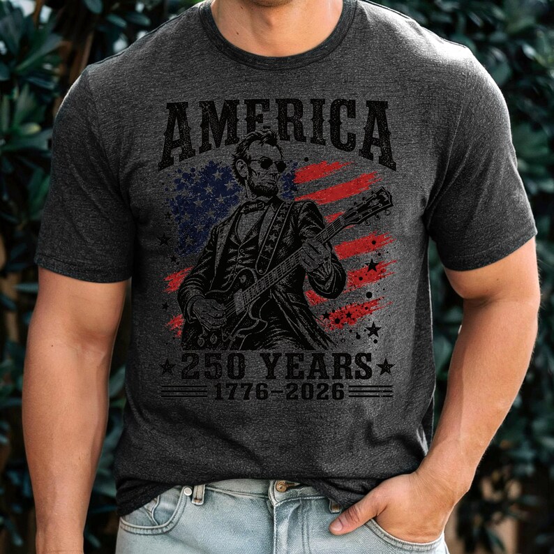 K&ouml;nnte beinhalten: Dunkelgraues T-Shirt mit einer Grafik von Abraham Lincoln, der Gitarre spielt, vor dem Hintergrund der amerikanischen Flagge. Oben steht "AMERICA", darunter "250 YEARS 1776-2026".