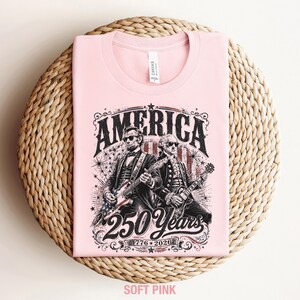 May include: A soft pink t-shirt featuring a graphic of two historical figures playing guitars, with the word "AMERICA" above them. The design includes the text "250 Years 1776-2026". The shirt is folded on a woven surface.