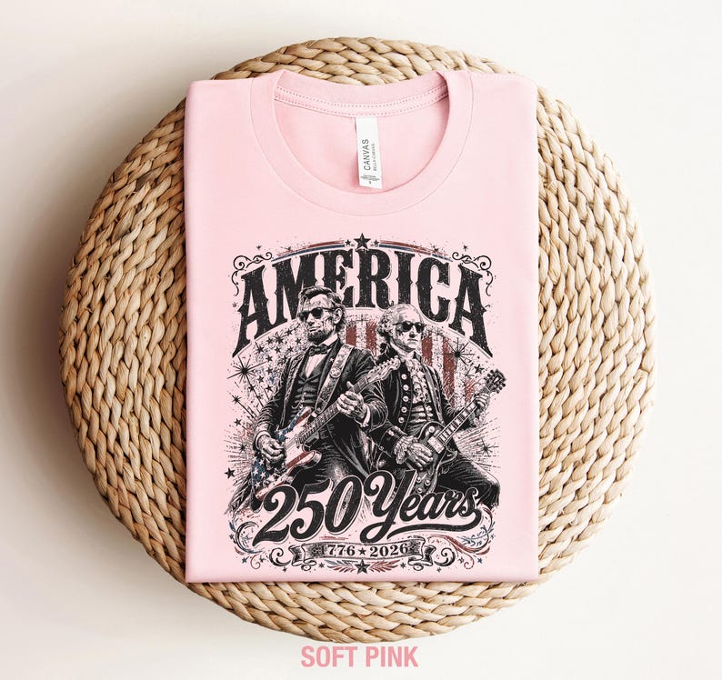 May include: A soft pink t-shirt featuring a graphic of two historical figures playing guitars, with the word "AMERICA" above them. The design includes the text "250 Years 1776-2026". The shirt is folded on a woven surface.