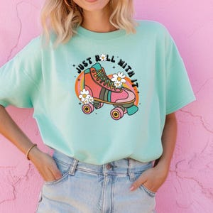 May include: A light mint green t-shirt featuring a graphic print of a pink and orange roller skate surrounded by white daisies. The text "JUST ROLL WITH IT" is arched above the skate in a black font, set against a circular rainbow background.