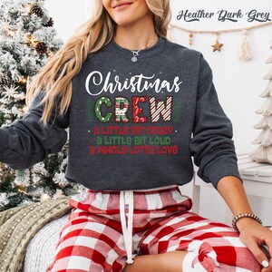 May include: Heather dark grey sweatshirt with the text "Christmas CREW" in white, red, and green. The sweatshirt also has the phrases "a little bit crazy", "a little bit loud", and "a whole lotta love". The person in the image is wearing red and white plaid pajama pants.