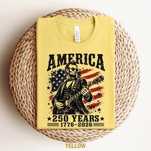 K&ouml;nnte beinhalten: Gelbes T-Shirt mit einer Grafik von Abraham Lincoln, der Gitarre vor einer amerikanischen Flagge spielt. Das Wort "AMERICA" steht &uuml;ber dem Bild, darunter "250 YEARS 1776-2026". Das T-Shirt wird auf einer gewebten Oberfl&auml;che pr&auml;sentiert.