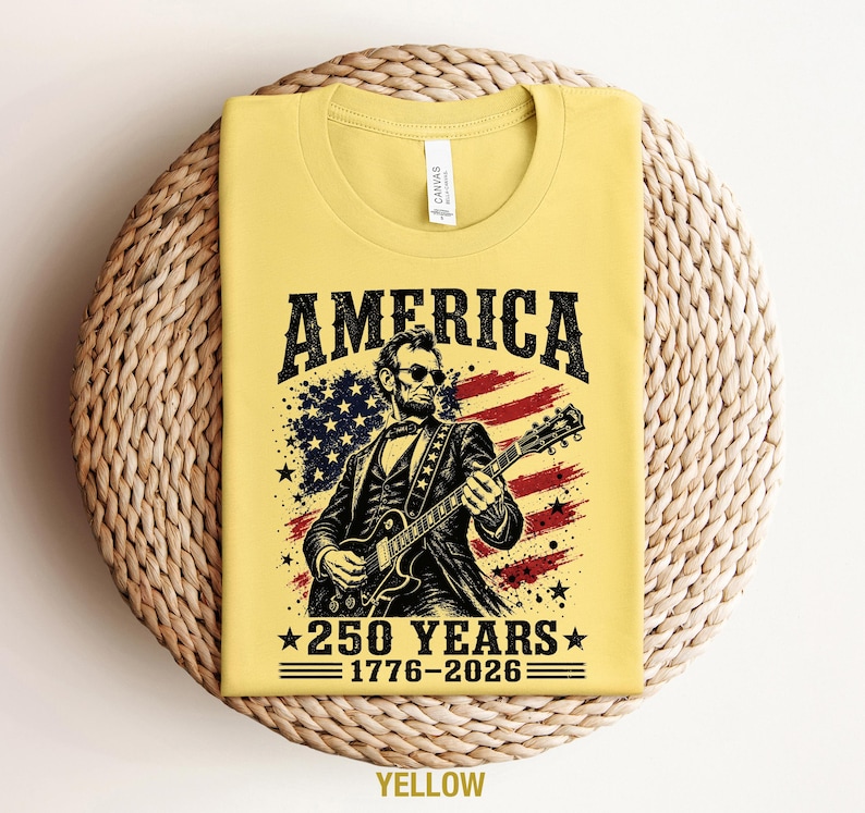 K&ouml;nnte beinhalten: Gelbes T-Shirt mit einer Grafik von Abraham Lincoln, der Gitarre vor einer amerikanischen Flagge spielt. Das Wort "AMERICA" steht &uuml;ber dem Bild, darunter "250 YEARS 1776-2026". Das T-Shirt wird auf einer gewebten Oberfl&auml;che pr&auml;sentiert.