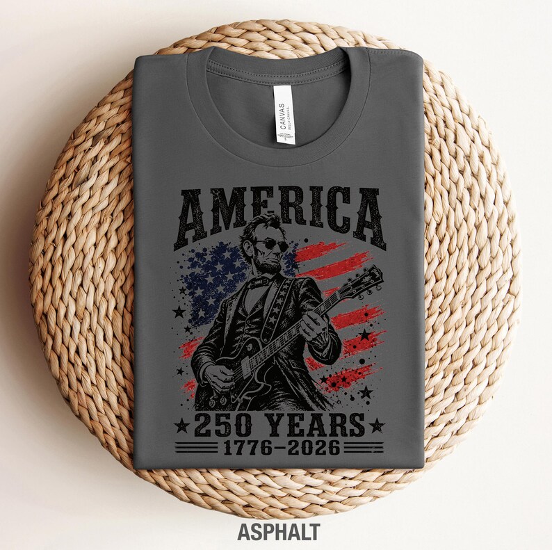 K&ouml;nnte beinhalten: Asphaltfarbenes T-Shirt mit einer Grafik von Abraham Lincoln, der vor einer amerikanischen Flagge Gitarre spielt. Der Text lautet "AMERICA" &uuml;ber dem Bild und "250 YEARS 1776-2026" darunter. Ein patriotisches Design.