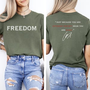 May include: Olive green t-shirt with the word "FREEDOM" printed in white on the front. The back of the shirt features the quote, "JUST BECAUSE YOU ARE OFFENDED DOESN'T MEAN YOU ARE RIGHT." in white and red, with a signature.