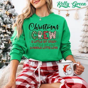 May include: A Kelly Green Christmas Crew sweatshirt with the text "Christmas Crew" in black and red. The sweatshirt also has the phrases "a little bit crazy", "a little bit loud", and "a whole lotta love". The person is wearing red and white plaid pajama pants.