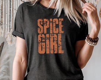 May include: Dark gray t-shirt with the words "SPICE GIRL" in distressed orange lettering. The shirt has short sleeves and a crew neck. The model is wearing blue jeans and a brown belt.