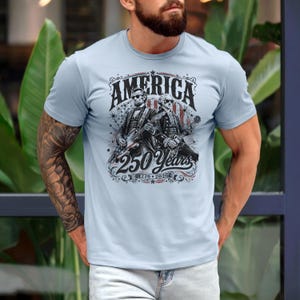 May include: Light blue t-shirt with a graphic featuring the word "AMERICA" above two figures playing guitars. The graphic also includes the text "250 Years" and the years "1776-2026". The design is in shades of black, white, and red.