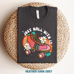May include: A heather dark grey t-shirt featuring a graphic of a pink and orange roller skate surrounded by white daisies and a circular rainbow background. The text "JUST ROLL WITH IT" is printed in a white arched font above the roller skate.