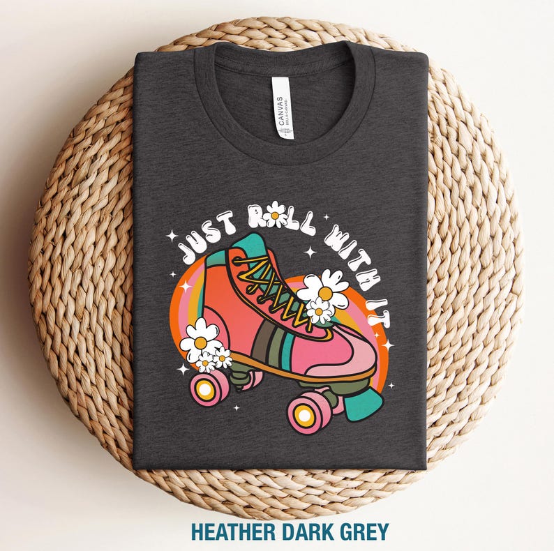 May include: A heather dark grey t-shirt featuring a graphic of a pink and orange roller skate surrounded by white daisies and a circular rainbow background. The text "JUST ROLL WITH IT" is printed in a white arched font above the roller skate.