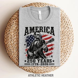 K&ouml;nnte beinhalten: Hellgraues T-Shirt mit einem Aufdruck von Abraham Lincoln, der Gitarre vor einer amerikanischen Flagge spielt. Der Text "AMERICA" und "250 YEARS 1776-2026" sind ebenfalls auf dem Shirt abgebildet. Das T-Shirt liegt gefaltet auf einer gewebten Oberfl&auml;che.