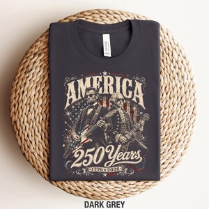 May include: Dark grey t-shirt featuring an illustration of Abraham Lincoln and George Washington playing guitars. The design includes the word "AMERICA" and "250 Years 1776-2026" in a vintage style.