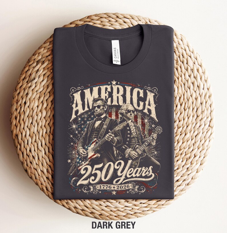 May include: Dark grey t-shirt featuring an illustration of Abraham Lincoln and George Washington playing guitars. The design includes the word "AMERICA" and "250 Years 1776-2026" in a vintage style.