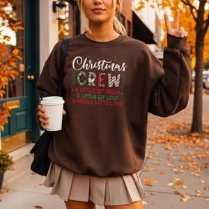 May include: A brown sweatshirt with the text "Christmas CREW" in white and green. The text below reads "a LITTLE BIT CRAZY. a LITTLE BIT LOUD a WHOLE LOTTA LOVE". The sweatshirt is a crew neck style.